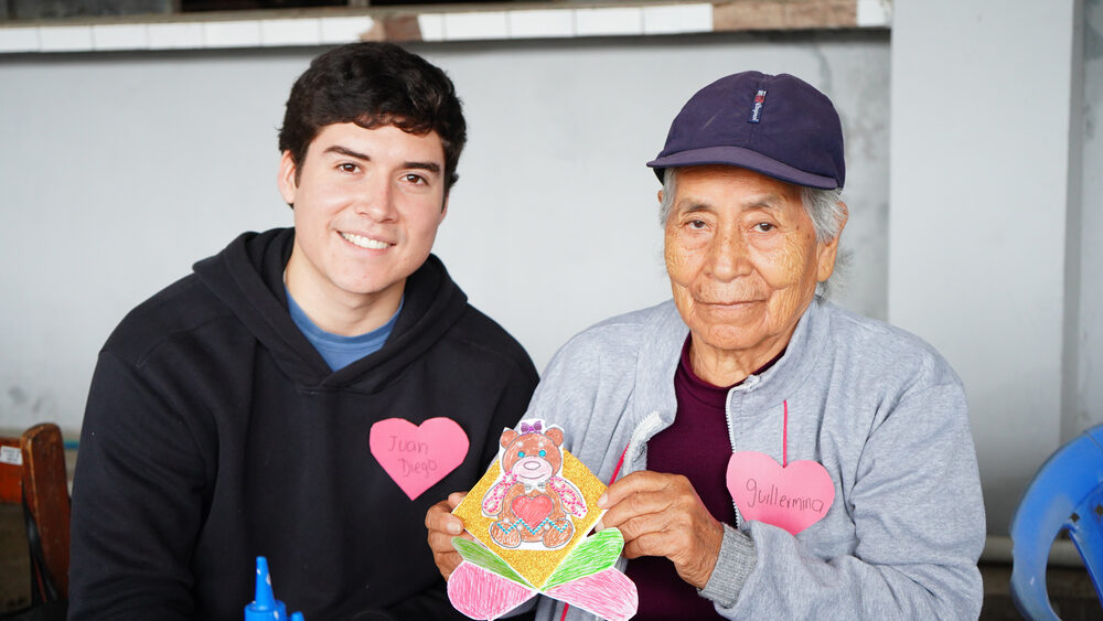 Voluntario joven abraza a adulto mayor con sombrero durante taller del programa