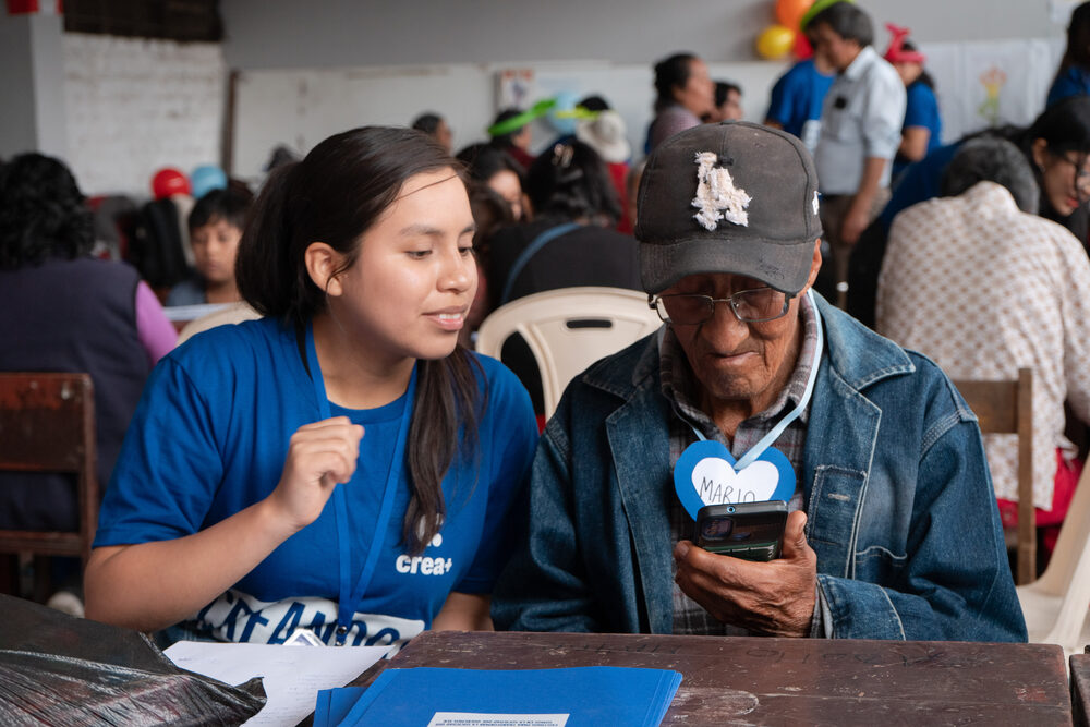 Voluntaria y adulto mayor ríen juntos mirando un celular en el taller