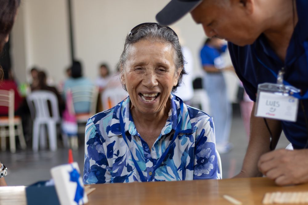 Adulta mayor riendo a carcajadas mientras voluntario joven a su lado escribe en el taller