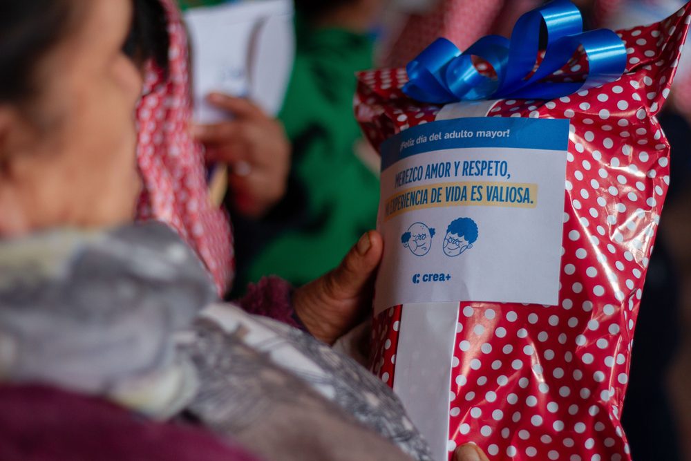 Detalle de manos de adulta mayor recibiendo bolsa con frase del programa